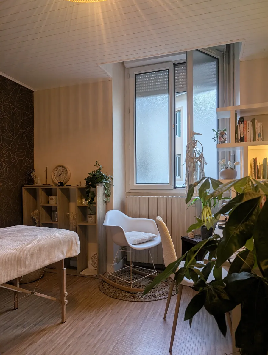 Le cabinet de Catherine Mercier, avec la table de soin, un fauteuil blanc près de la fenêtre et des plantes vertes